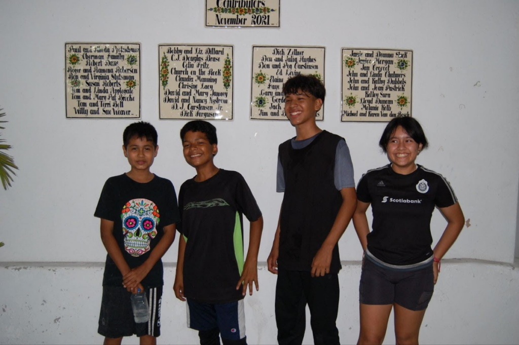 Four young boys standing in front of a wall with names of donors, reflecting a sense of community involvement.