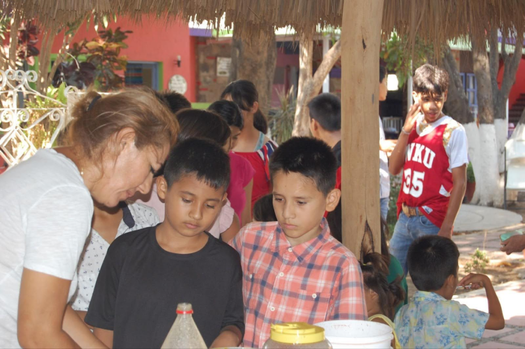 Two boys engaged in an activity with an adult, while others stand around in the background, showing participation in a group setting.