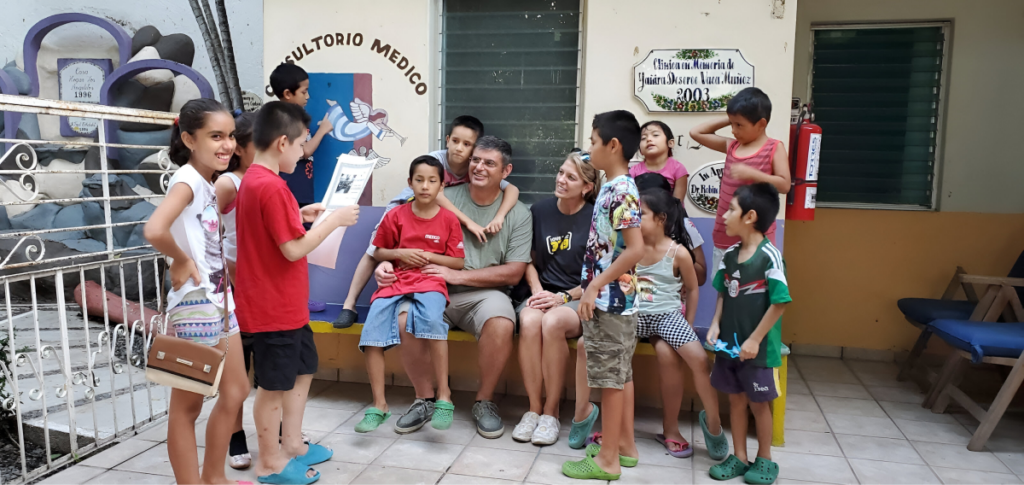 Children gathered around two adults outdoors, one child reading from a paper.